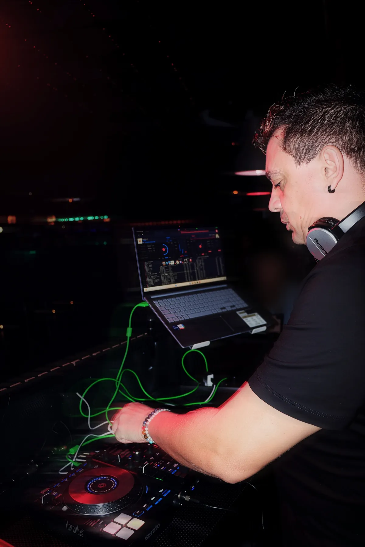 DANKO DJ de perfil en la cabina, con auriculares al cuello, mezclando con su portátil y su controladora